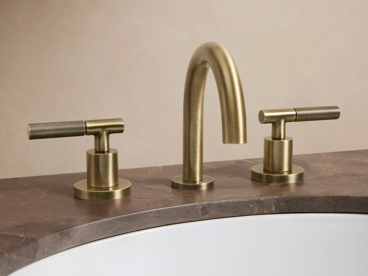 Crewe Knurled Faucet in antique brass, featuring a curved spout and two cylindrical knurled handles mounted on a marble countertop.