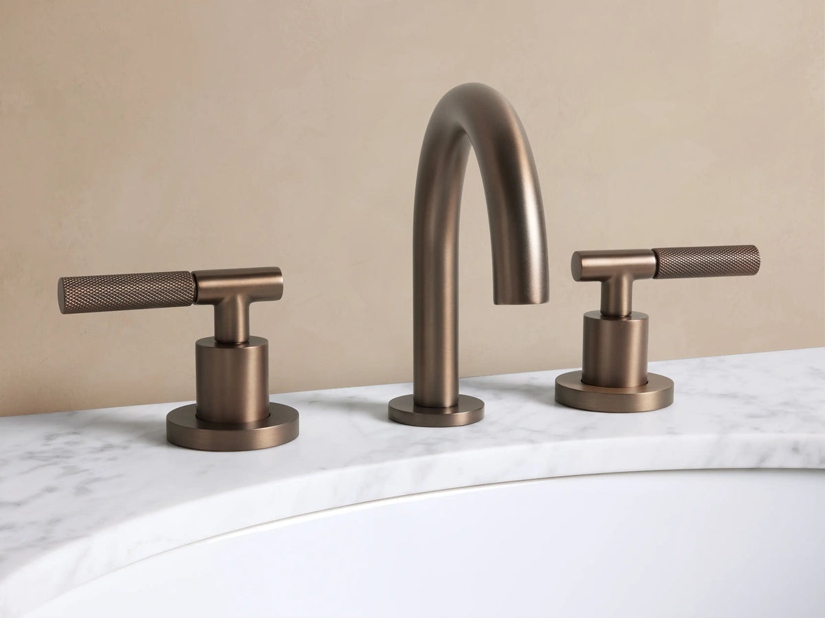 Crewe Knurled Faucet in Bronze: Three-piece widespread bronze faucet with a gooseneck spout & knurled T-handles on white marble.