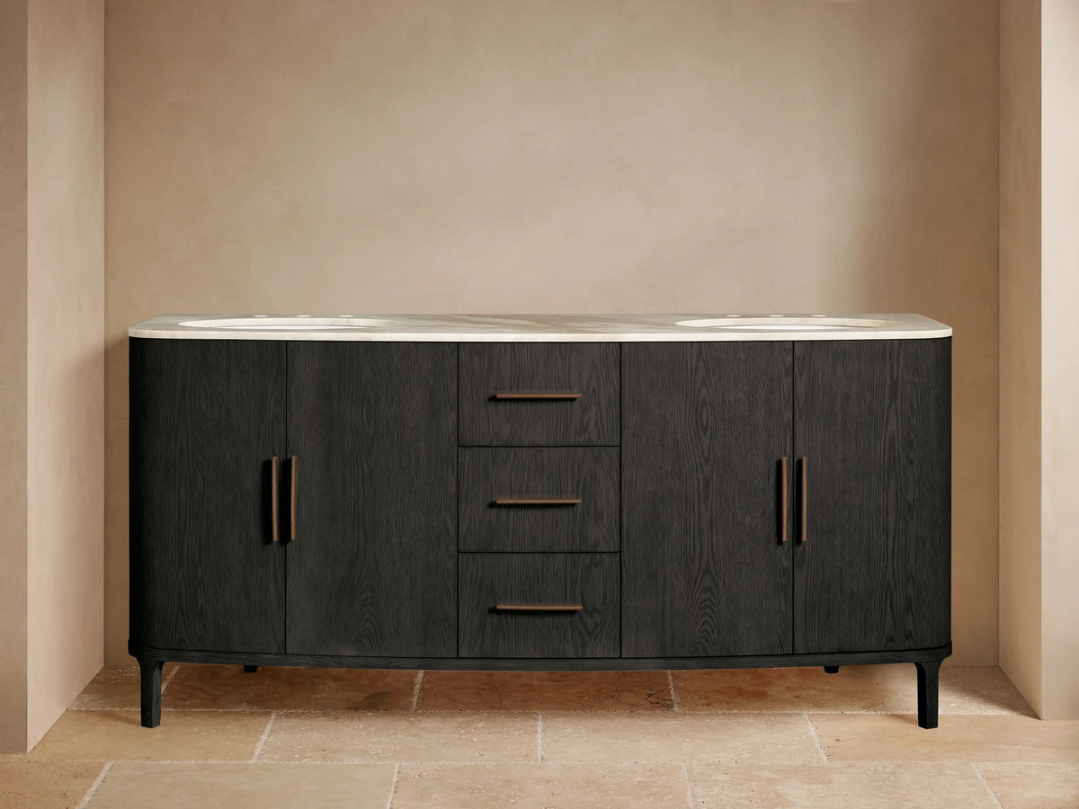 Danny Double Vanity, dark oak with curved ends & light marble top. Two sinks, bronze faucets, left door ajar with towels.