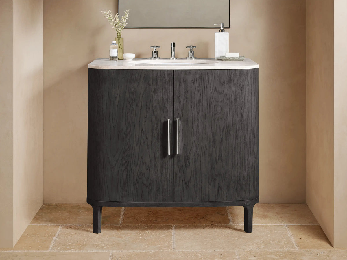 Danny Single Vanity in dark oak with rounded edges, a white stone top, two metallic handles, and a ceramic sink, set against a beige tile wall.