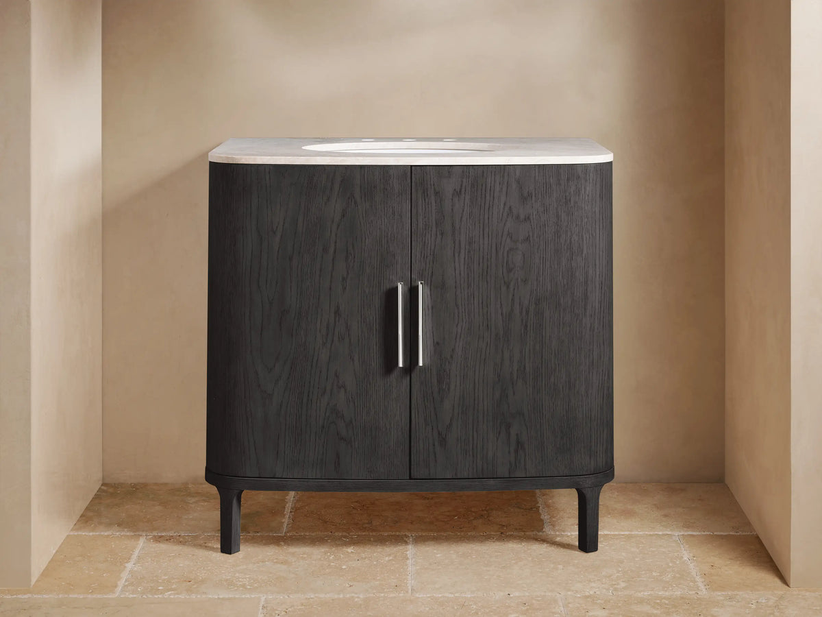 Danny Single Vanity in dark oak with rounded edges, a white stone top, two metallic handles, and a ceramic sink, set against a beige tile wall.