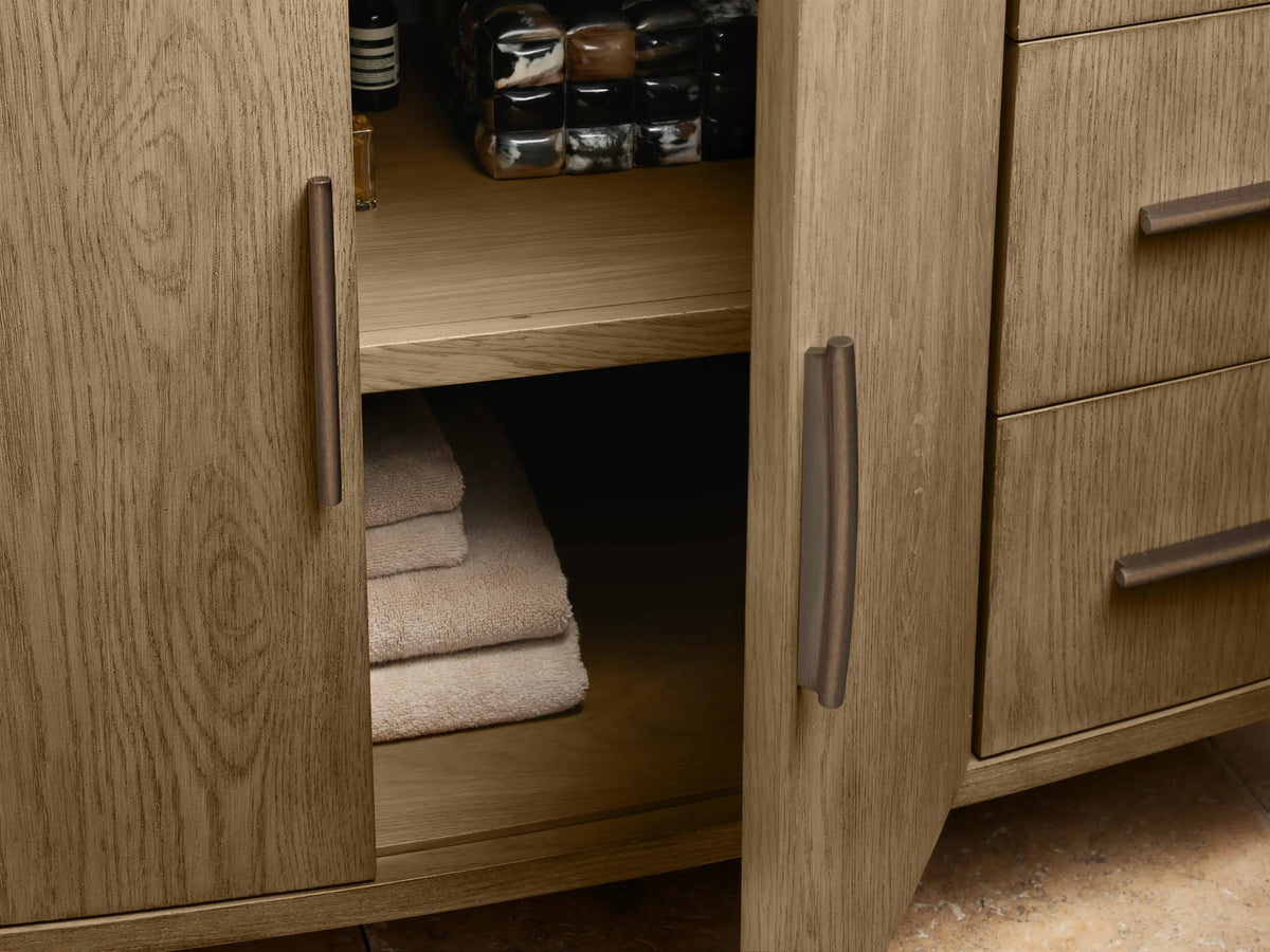 Close-up of Danny Wide Single Vanity. Light oak vanity with white marble top, dark bronze handles. Bottom drawer slightly open, revealing white towel.