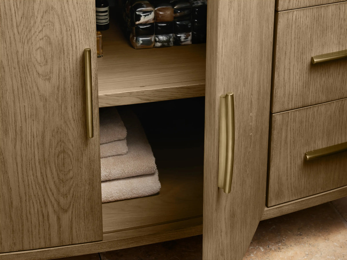 Oak wood Danny Single Wide Vanity with curved front design, featuring open drawer revealing a folded towel, gold-colored handles, and marble top edge.