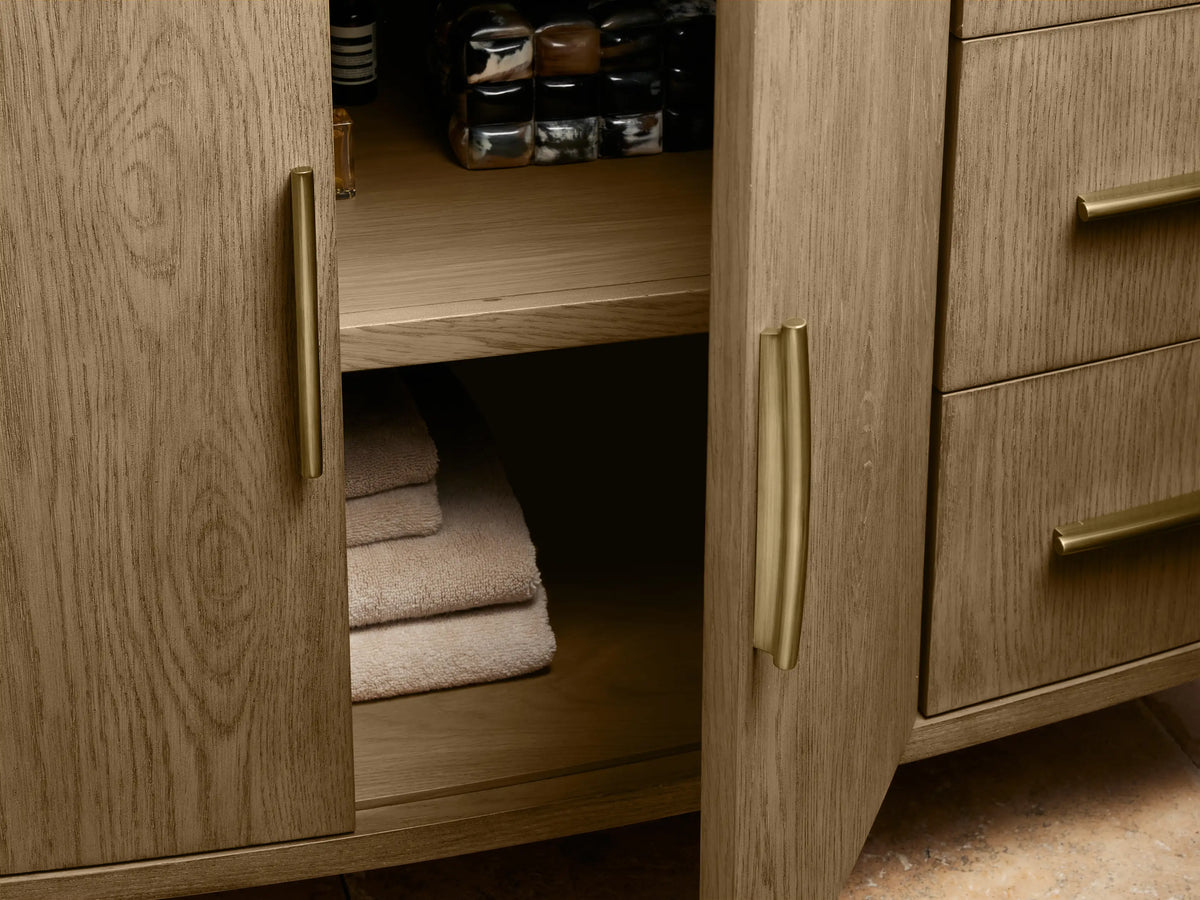 Danny Single Wide Vanity with curved oak wood finish, open drawer revealing a towel, and brass hardware on soft-close glides.