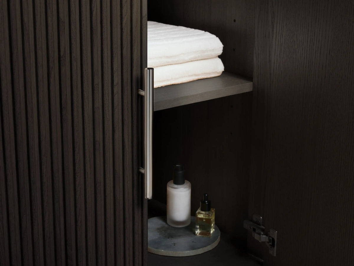 Partially open drawer of Finnley Double Vanity, dark fluted wood, polished nickel handle, with white towels and bottle inside. Dark countertop.