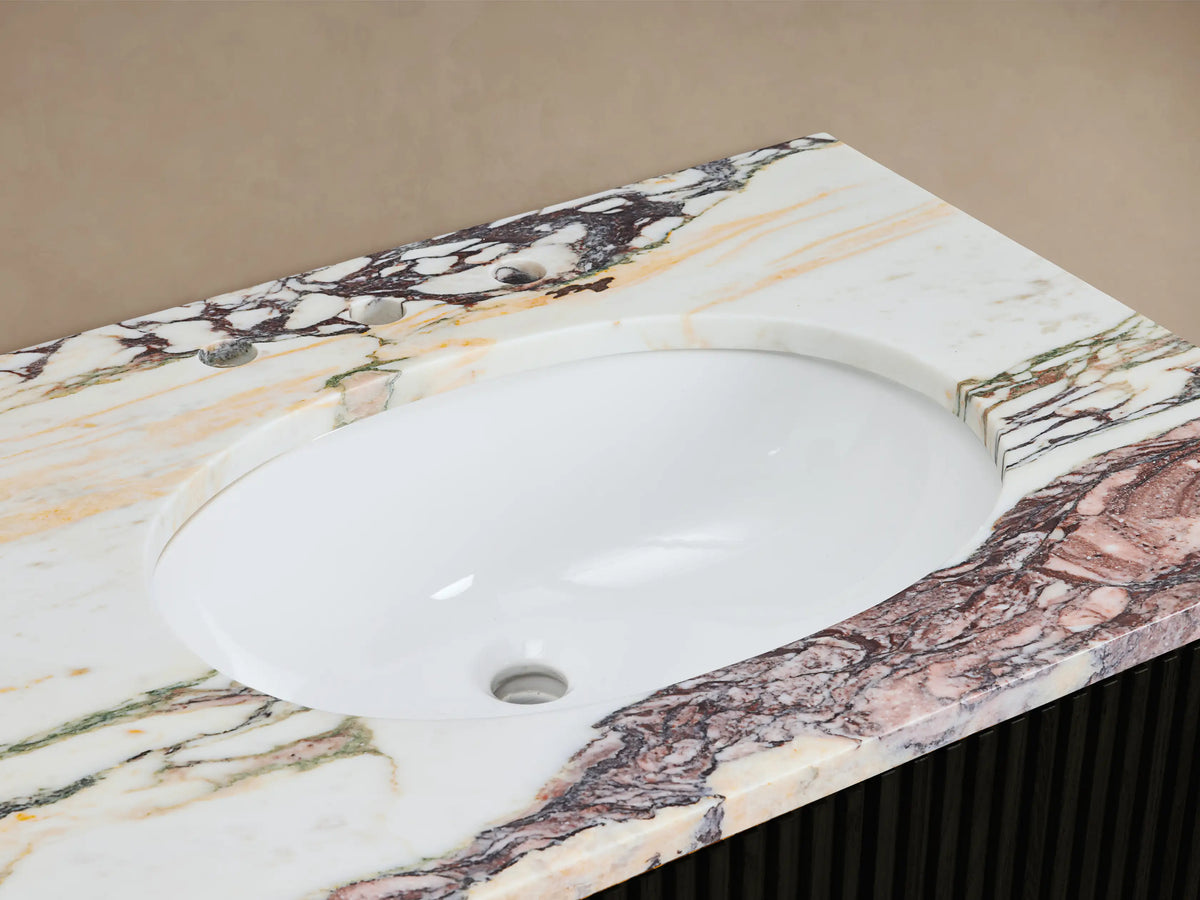 Close-up Finnley Vanity sink: white oval basin, antique brass faucet on multi-color veined marble countertop, with soap dispenser & towel.