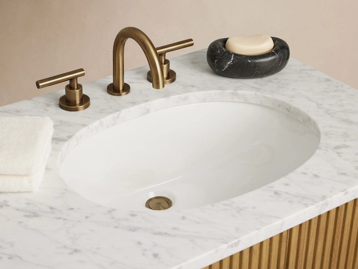 Finnley Double Vanity with ribbed oak wood finish, dual oval ceramic sinks, marble top, and antique brass hardware in a minimalist setting.
