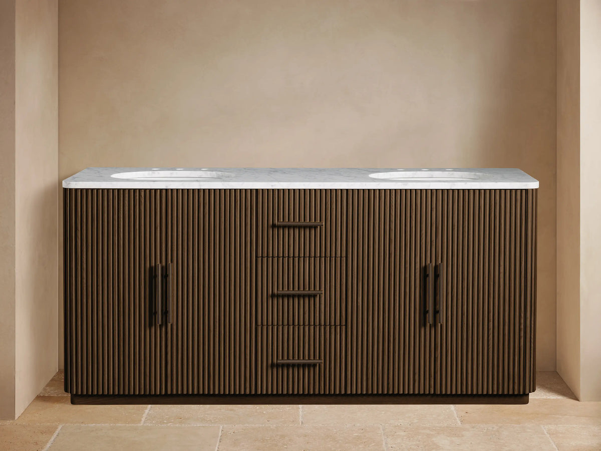 Finnley Double Vanity in dark fluted wood with white marble top, two sinks, bronze faucets. Left cabinet door open revealing towels.