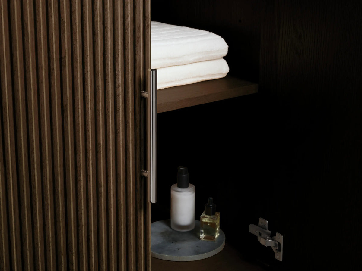 Finnley Double Vanity close-up. Ribbed dark oak drawer slightly open with white towels & bottle. White marble top, silver handle.