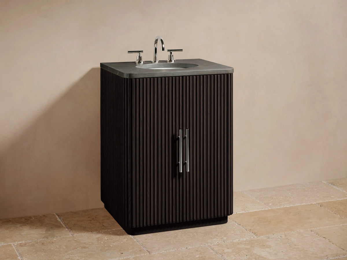 Alt text: Finnley Powder Vanity with a dark wood finish, vertical grooves, stone countertop, and chrome faucet in a beige setting.