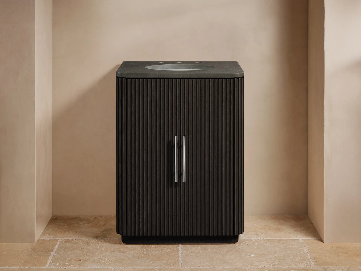 Dark oak wood vanity cabinet door partially open, revealing shelves with neatly folded towels and bottles. Silver vertical handle on ribbed texture.