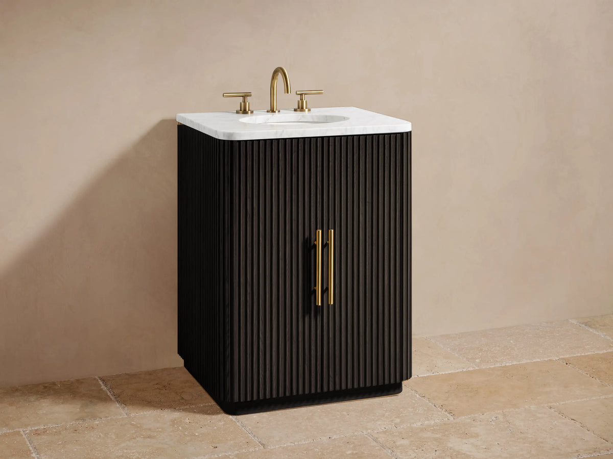 Finnley Powder Vanity with ribbed dark wood door partially open, revealing shelves with a round canister, bottle, and folded towels. Antique brass handle.