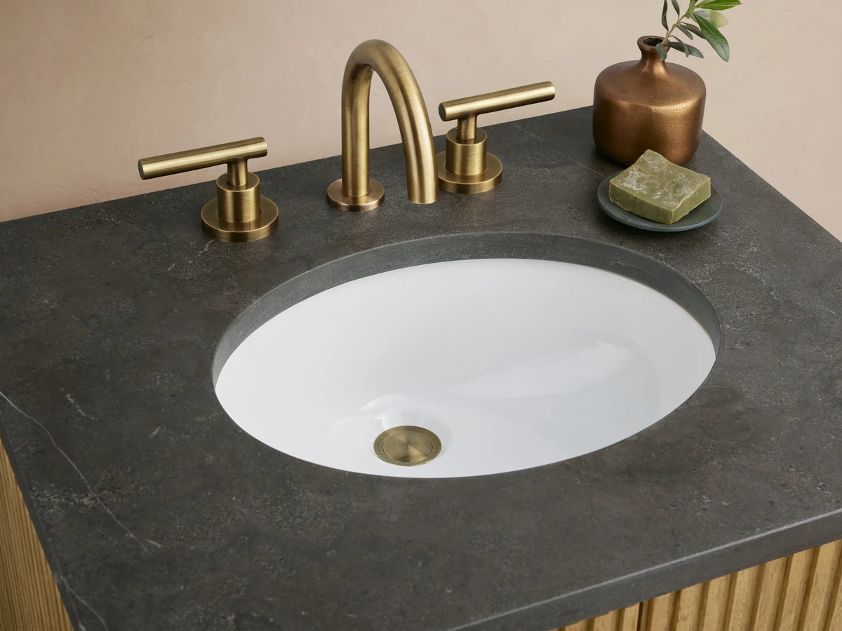 Finnley Powder Vanity with oak wood slatted front, black stone top, oval sink, and dual handle brass faucets, against a beige backdrop.