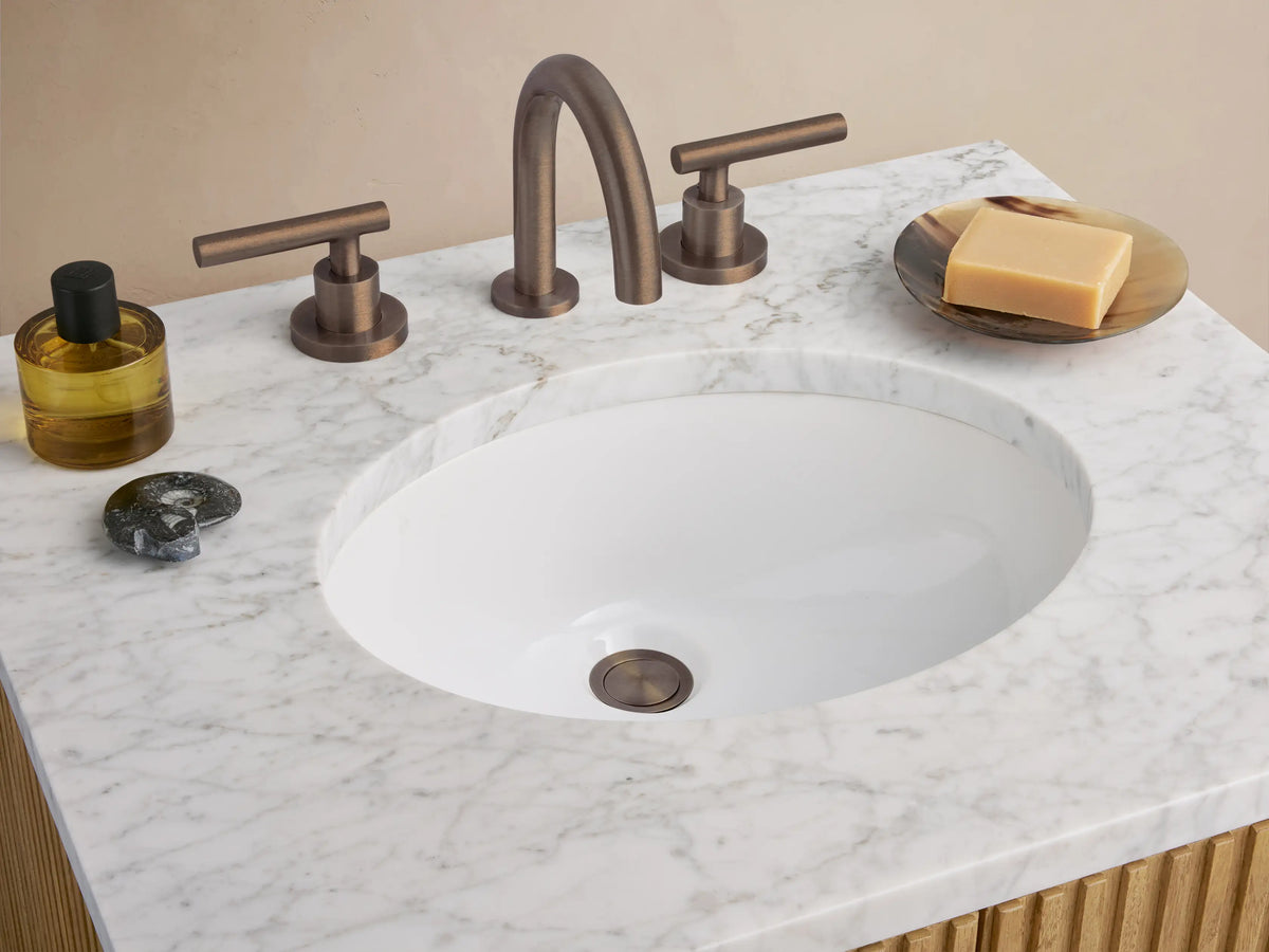 Finnley Powder Vanity: light oak cabinet, vertical fluting, dark handles, white marble top, oval sink, against a beige wall.