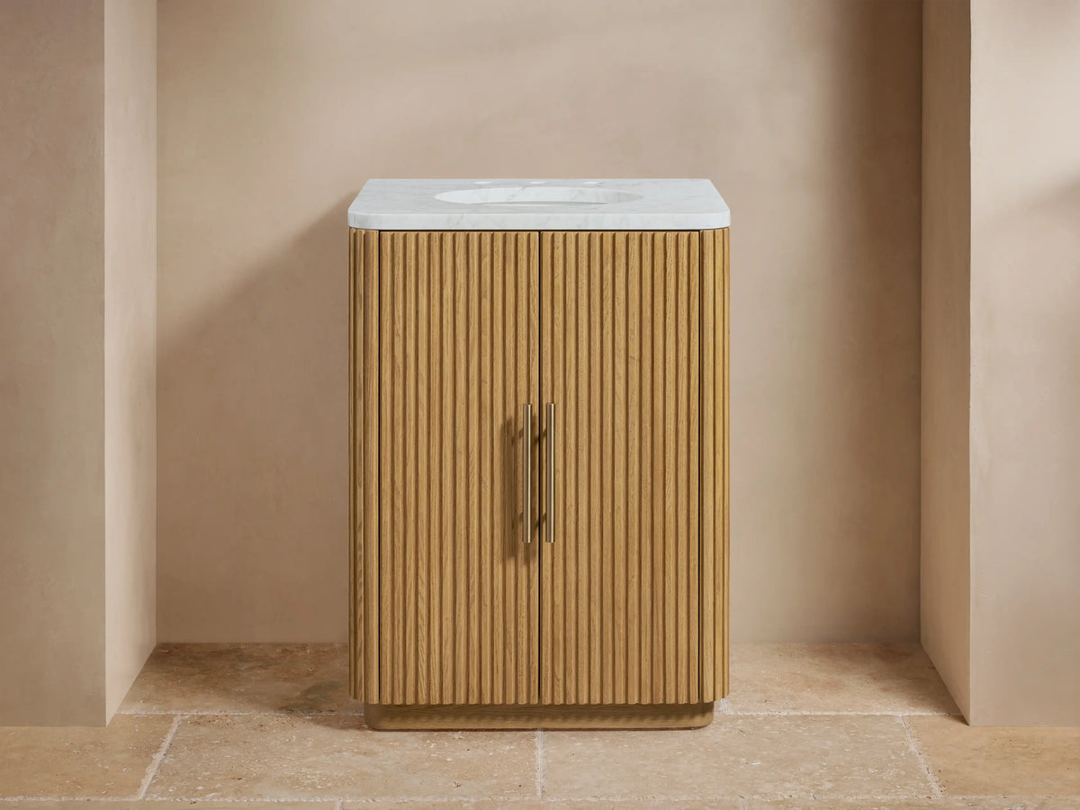 Oak wood vanity with vertical slat design, stone top, oval ceramic sink, and two brass-finished handles; beige backdrop.