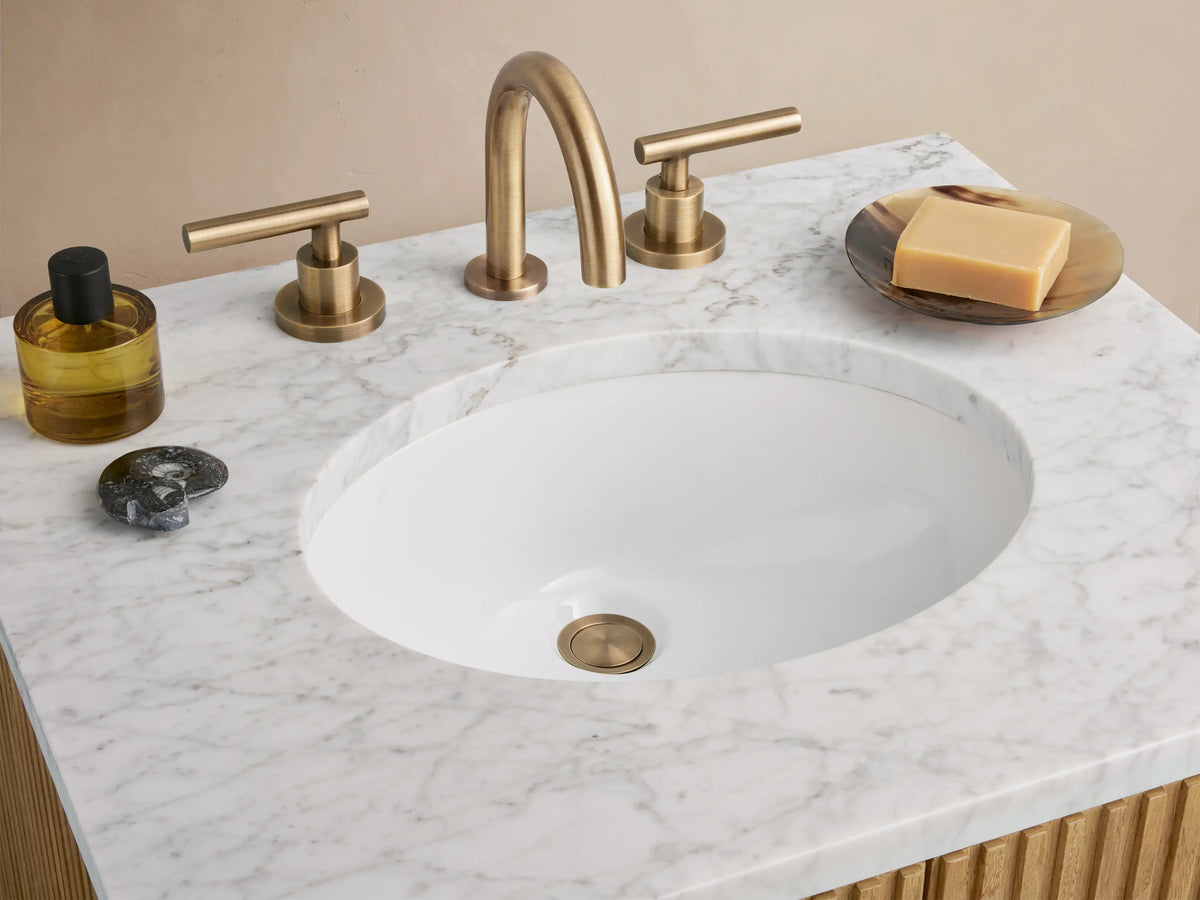 Finnley Powder Vanity with oak wood slatted front, brass handle, and white stone top. Sits on tile with beige wall backdrop.