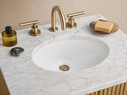 Finnley Powder Vanity with oak wood slatted front, brass handle, and white stone top. Sits on tile with beige wall backdrop.
