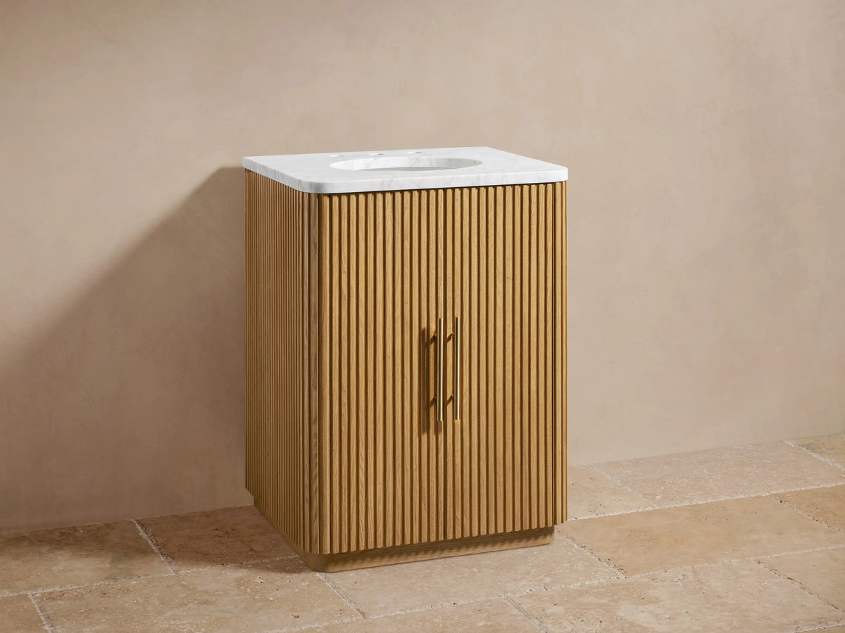 A wooden vanity with vertical slats, featuring a white stone top and oval sink. Brass faucets and handles are visible in front.