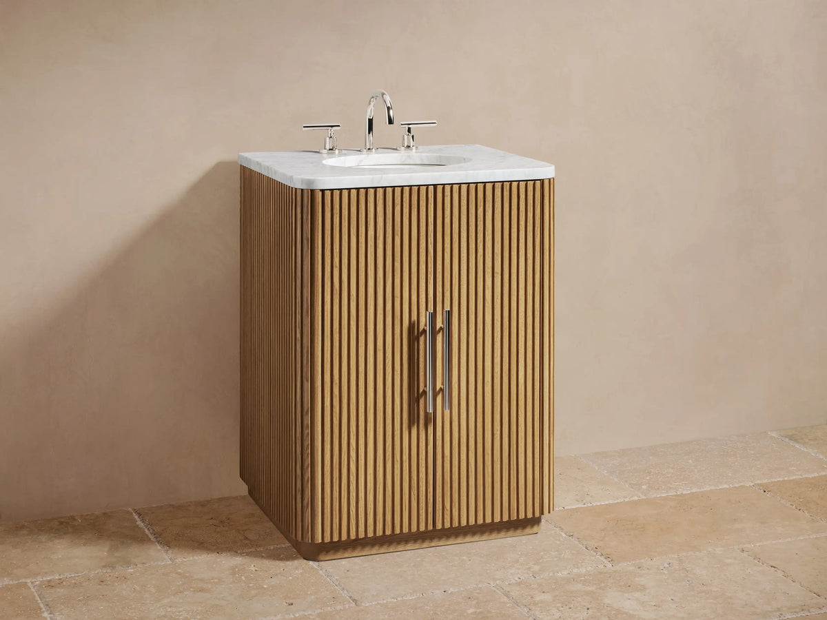 Open oak cabinet with vertical grooves, showing shelves containing folded beige towels and a small jar, metal handle on door.