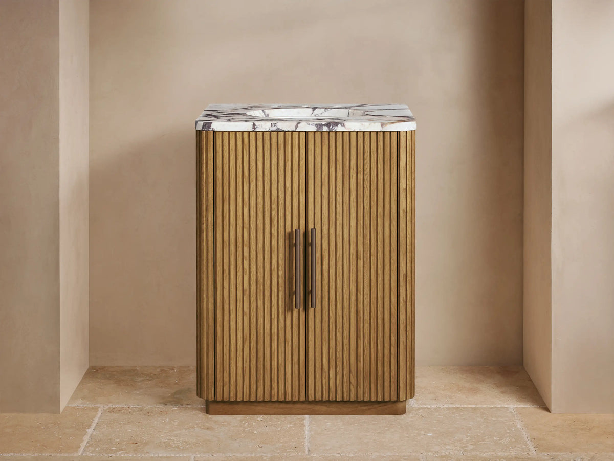 Finnley Powder Vanity: Ribbed oak cabinet, white marble top with bronze faucet & handles, against a beige wall on stone floor.