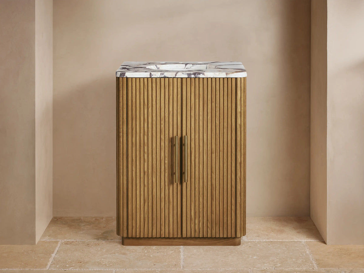 Finnley Powder Vanity: Light fluted wood cabinet, white marble top with brass faucet, soap dispenser, and small vase.