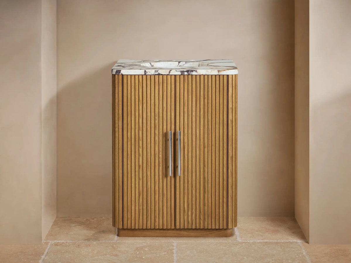 Finnley Powder Vanity: Fluted oak wood cabinet, white Carrara marble top, chrome faucet & handles. Beige wall, tiled floor.