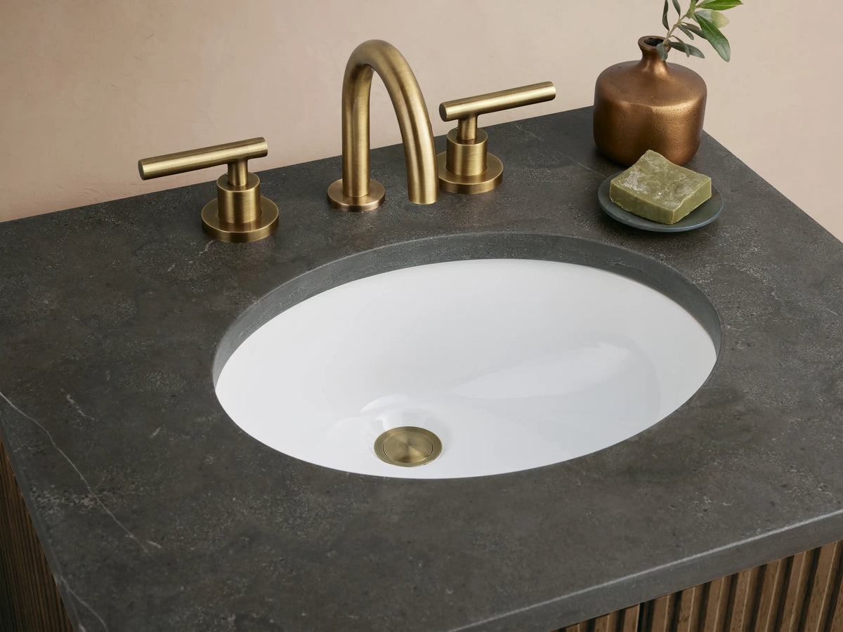 Finnley Powder Vanity: dark ribbed wood cabinet, brass handles, dark stone top, and oval sink, in a light room.