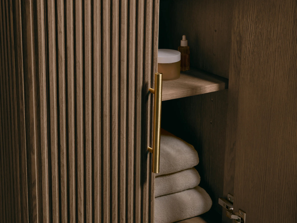 Finnley Powder Vanity with dark gray honed bluestone top, white oval undermount sink, brass widespread faucet. Slatted wood base.