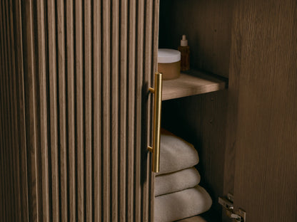 Finnley Powder Vanity with dark gray honed bluestone top, white oval undermount sink, brass widespread faucet. Slatted wood base.