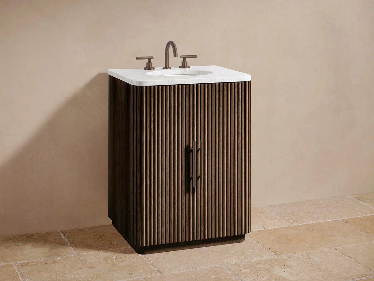 Finnley Powder Vanity cabinet, fluted light wood door ajar with brushed nickel handle, revealing cosmetics on shelf, stacked towels.
