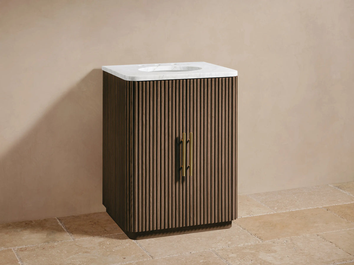 Finnley Powder Vanity: dark brown reeded wood, white marble top, oval sink, brass faucet/handles. Beige wall, tiled floor.