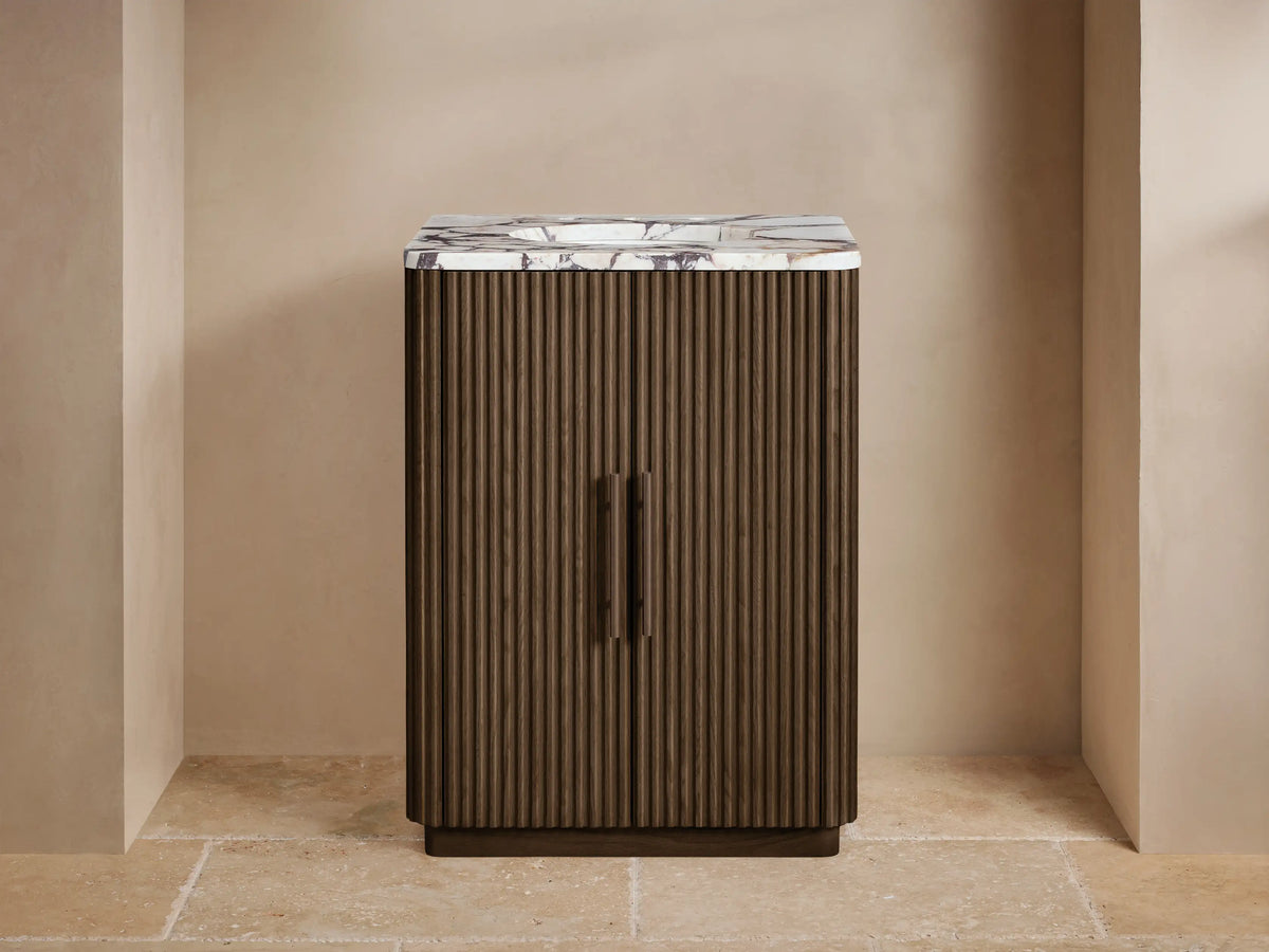 Finnley Powder Vanity: Dark reeded wood, white marble top, bronze faucet, vase, & soap dispenser in a beige room.