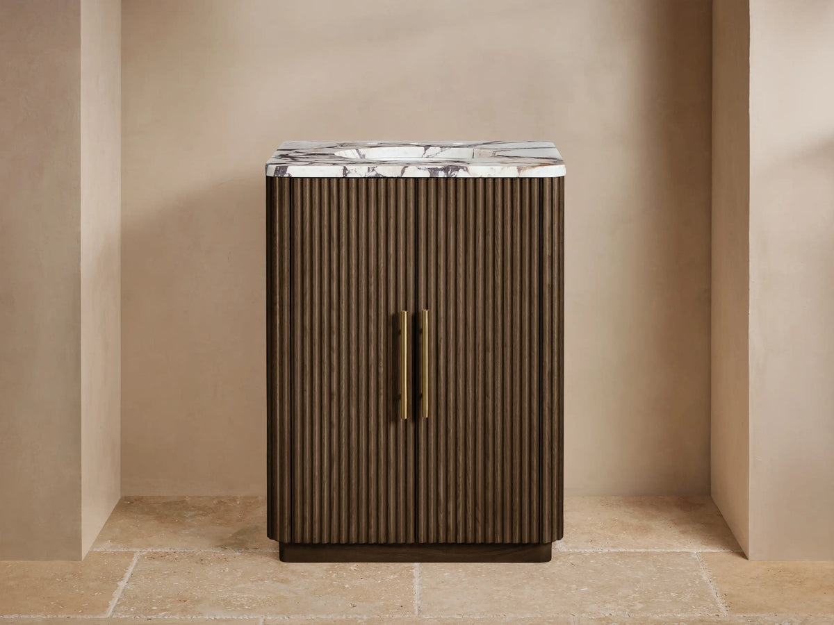 Finnley Powder Vanity: dark fluted wood cabinet, white marble top, gold faucet, plant, on stone floor.