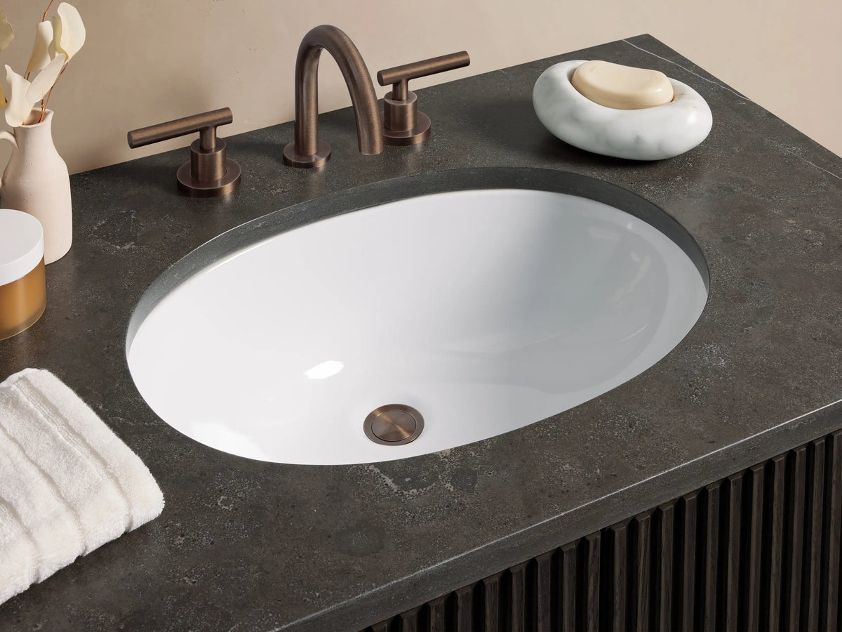 Finnley Single Vanity: dark brown ribbed cabinet, bronze handles, gray stone top with white oval sink, in a beige bathroom.