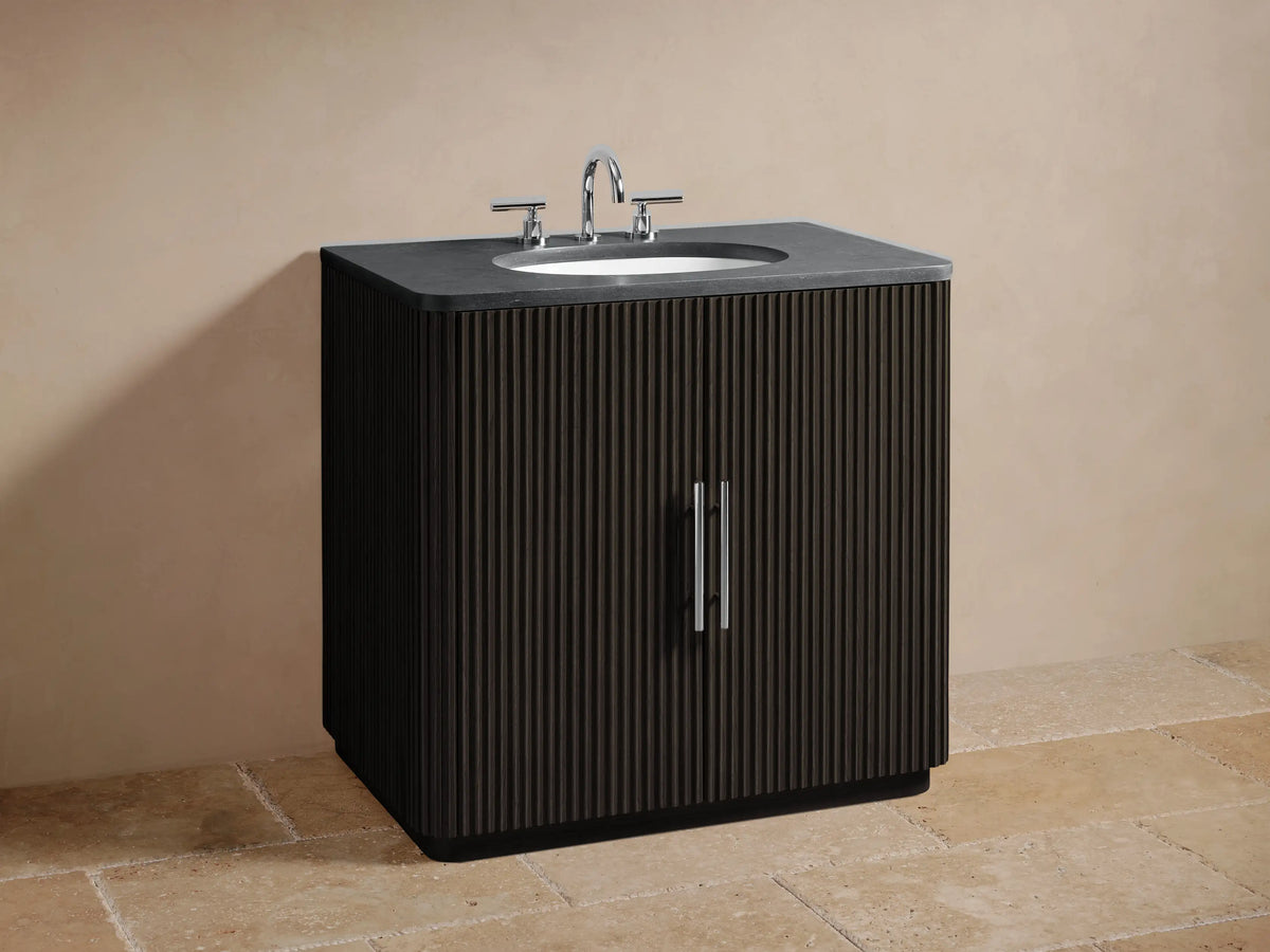 Dark wood vanity with vertical grooves, black stone top with oval sink. Two central handles on cabinet doors, in a neutral-toned environment.