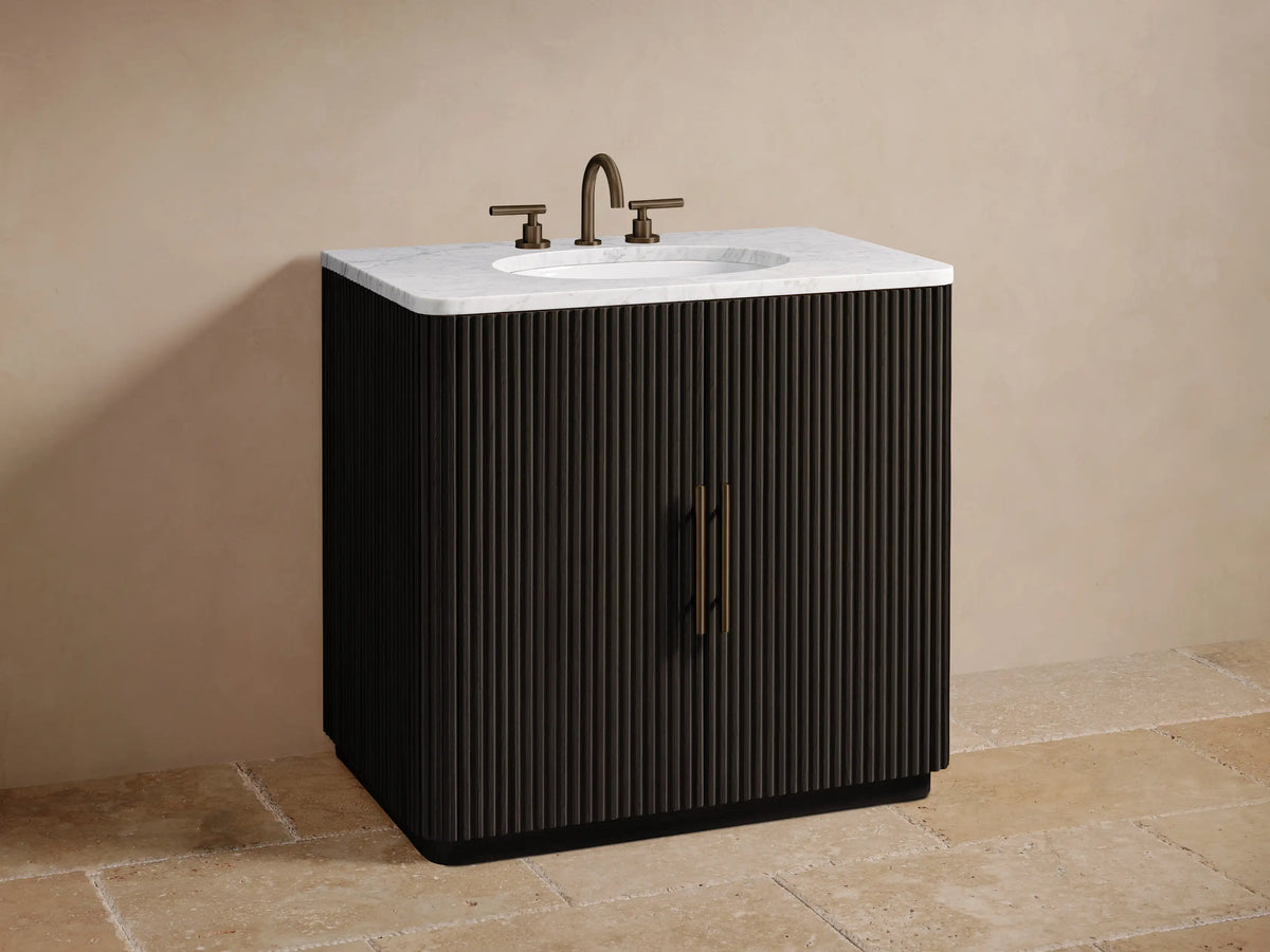 Finnley Single Vanity's open dark fluted cabinet door with a brass handle, showing interior shelf with a wooden tray & towels.