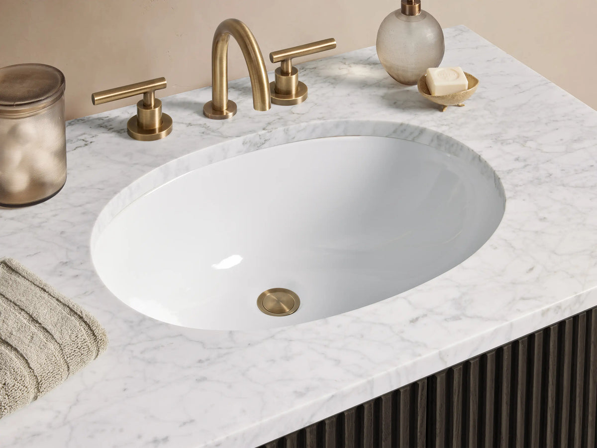 Finnley Single Vanity with dark oak slatted front, brass handles, and white stone top with oval sink, set against beige surroundings.