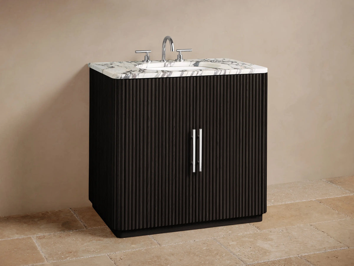Finnley Single Vanity with dark ribbed door, polished nickel handle, white Carrara marble top. Inside, toiletries and cream towels.