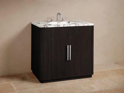 Finnley Single Vanity with dark ribbed door, polished nickel handle, white Carrara marble top. Inside, toiletries and cream towels.
