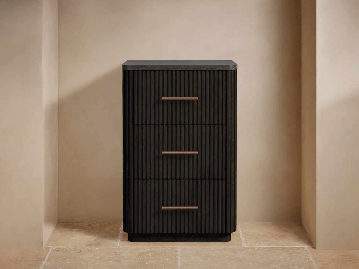 Finnley Short Bath Cabinet, dark ribbed wood, 3 drawers with bronze pulls, black stone top, towels & vase. Beige wall, stone floor.