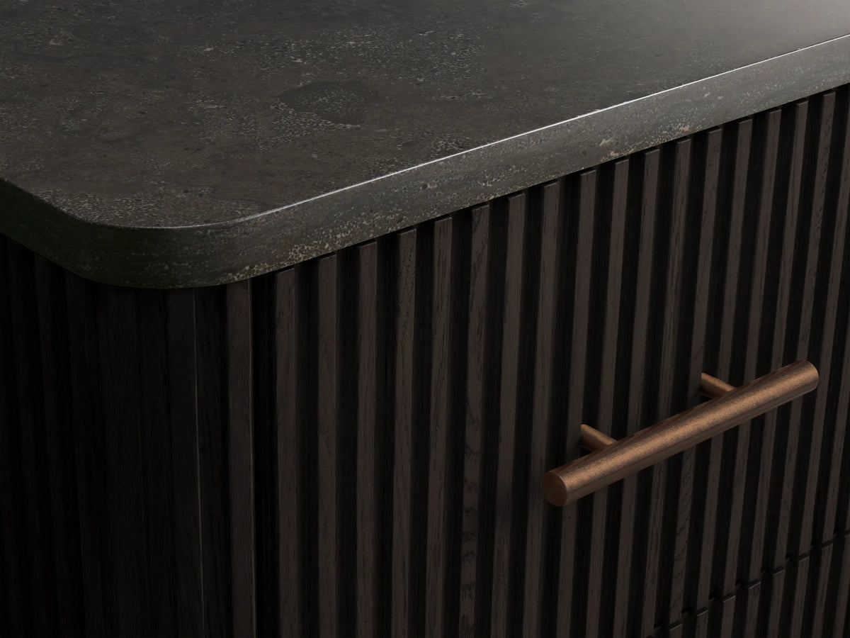 Front view of a dark, ribbed Finnley Short Bath Cabinet featuring a dark stone top and three drawers with antique bronze pull handles.