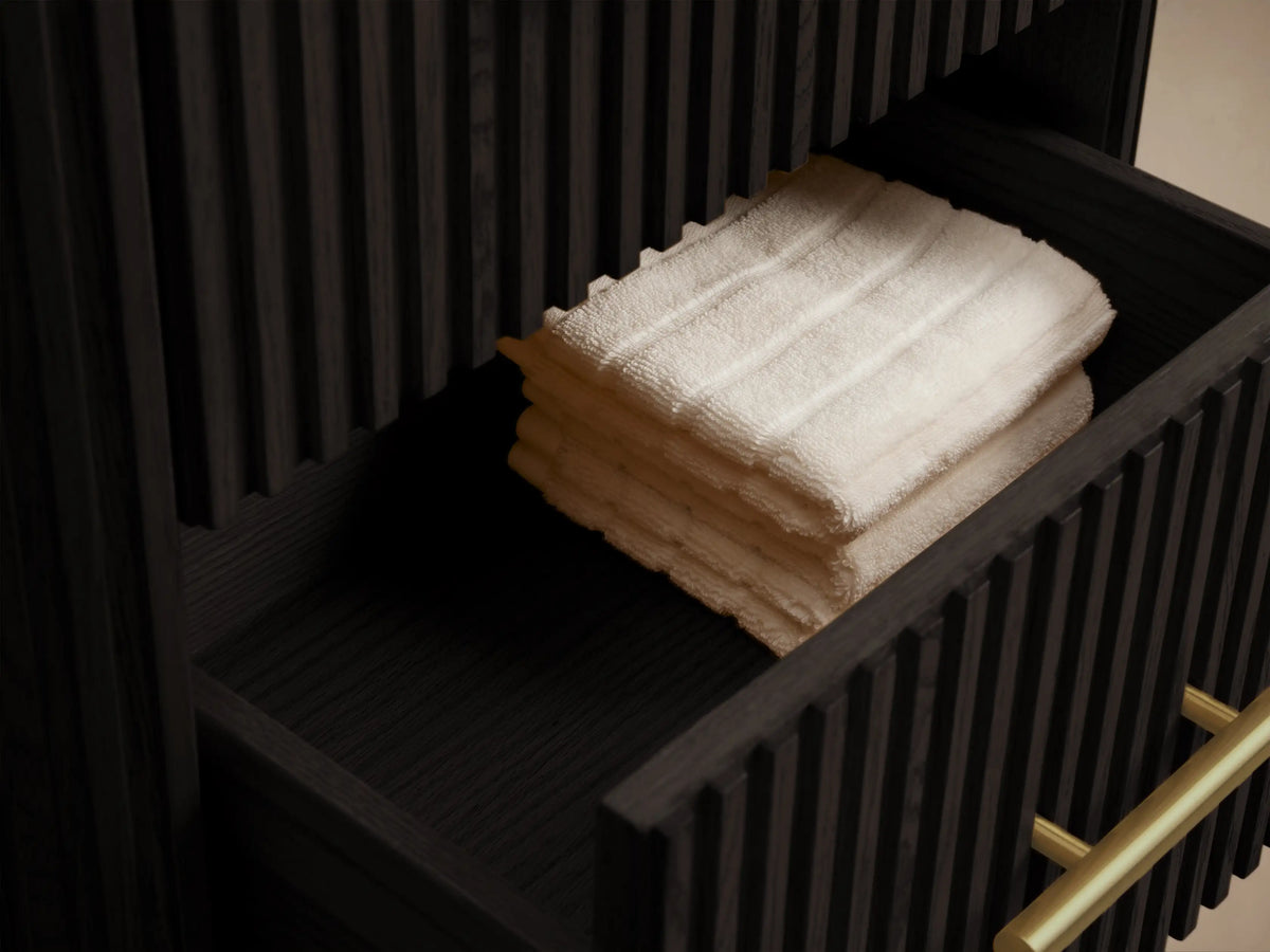 Finnley Short Bath Cabinet: Dark oak, fluted cabinet with three drawers, gold handles, and a dark stone top against a beige wall.