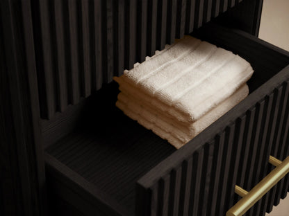 Finnley Short Bath Cabinet: Dark oak, fluted cabinet with three drawers, gold handles, and a dark stone top against a beige wall.