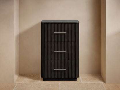 Finnley Short Bath Cabinet, dark ribbed wood, 3 drawers with silver pulls, dark stone top. Grey towels & flowers on top.