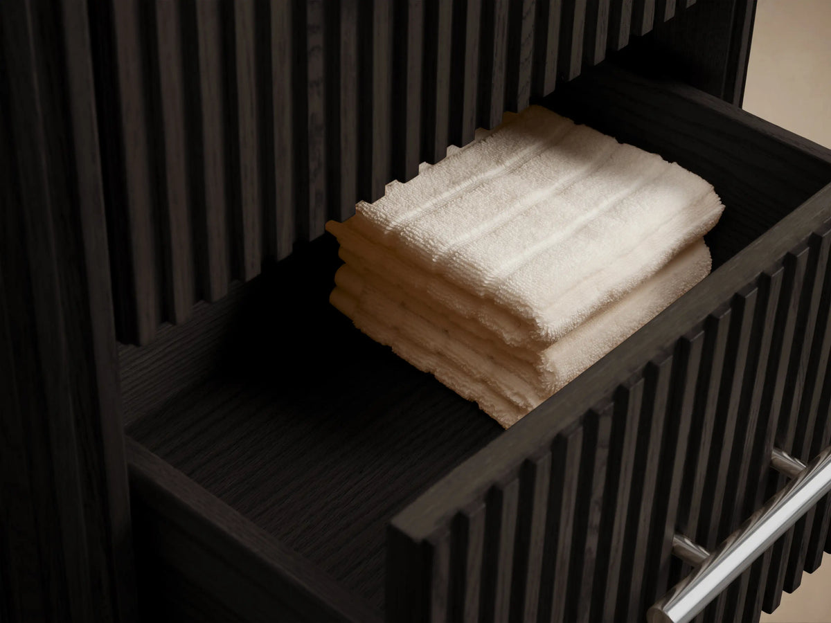 Dark oak Finnley Short Bath Cabinet with fluted details, 3 drawers, silver pulls & dark stone top, against a beige wall.