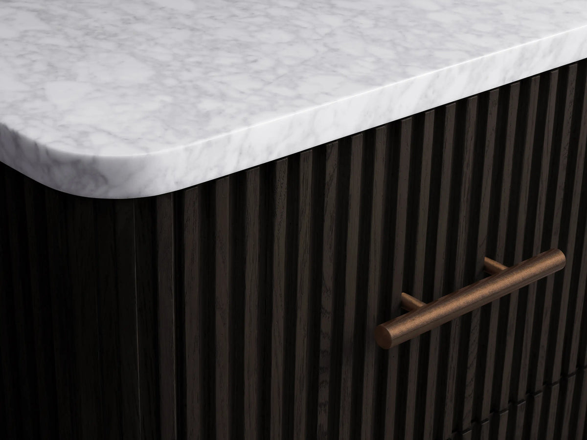 Finnley Short Bath Cabinet. Dark ribbed cabinet with white marble top, 3 drawers, antique brass pulls, on stone floor.