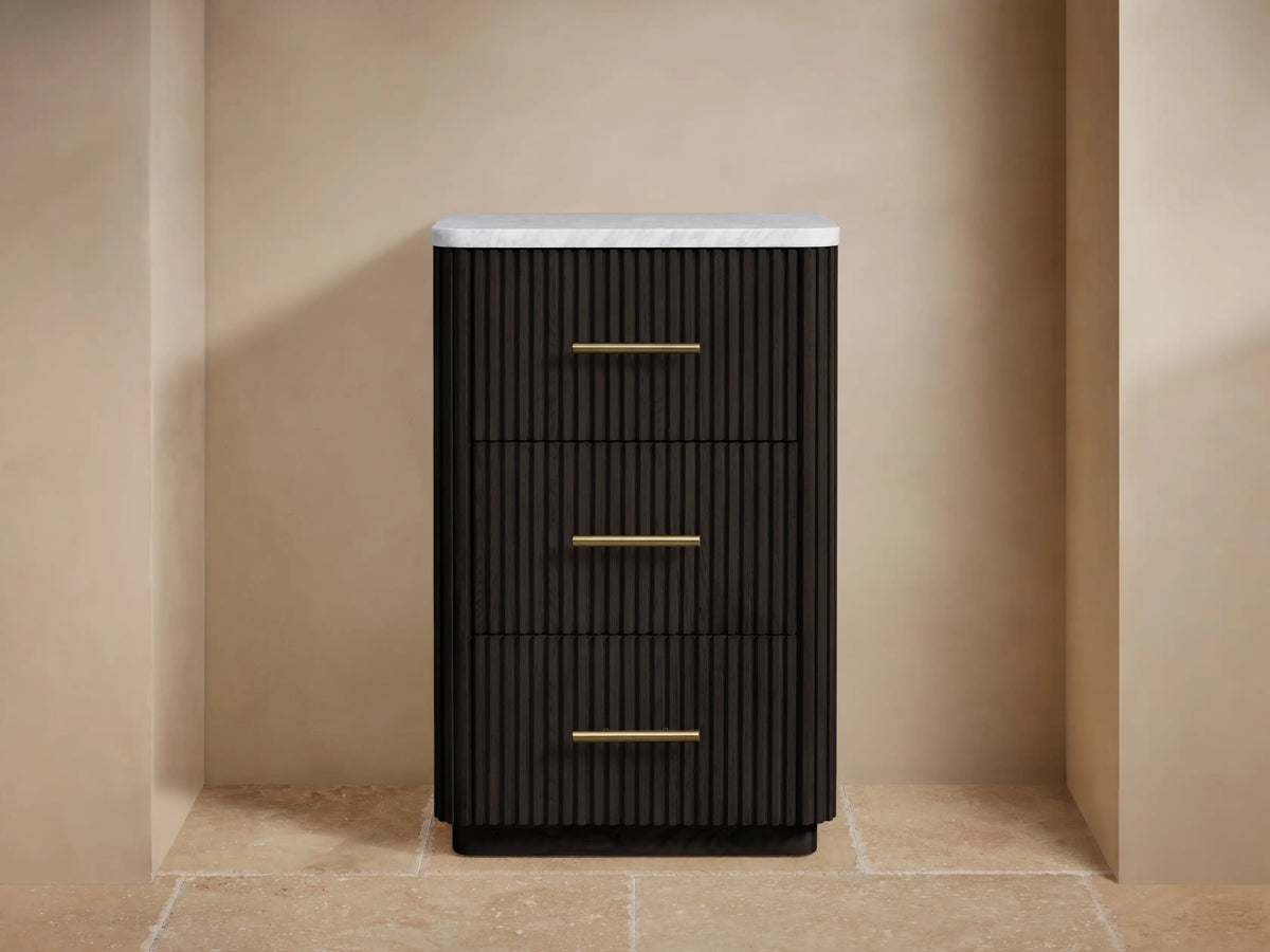 Finnley Short Bath Cabinet: dark fluted wood, white marble top, 3 drawers with brass pulls, beige wall, stone floor.