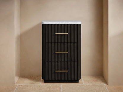 Finnley Short Bath Cabinet: dark fluted wood, white marble top, 3 drawers with brass pulls, beige wall, stone floor.