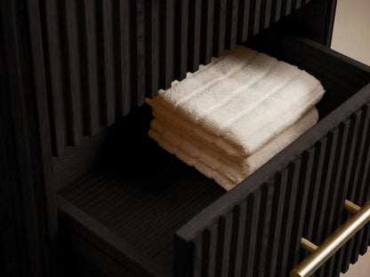 Finnley Short Bath Cabinet: Dark fluted wood cabinet with white marble top, 3 gold bar handles, against a beige wall & stone floor.