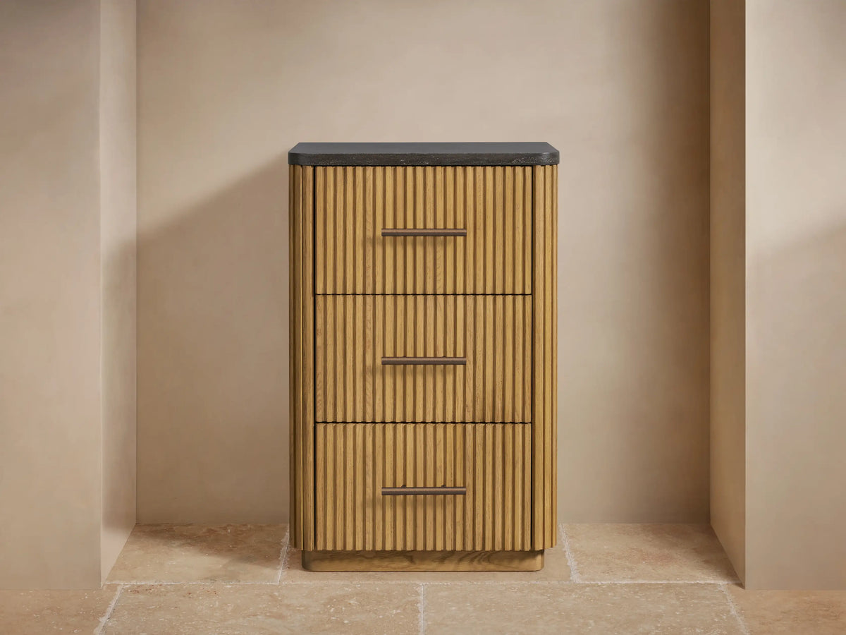 Finnley Short Bath Cabinet: light oak fluted wood, dark stone top, 3 drawers with bronze pulls, on tan wall and stone floor.